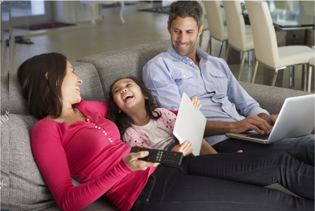 Family laughing while sitting on the sofa with the child on a tablet device, the father on a lap top, and mother holding television remote.